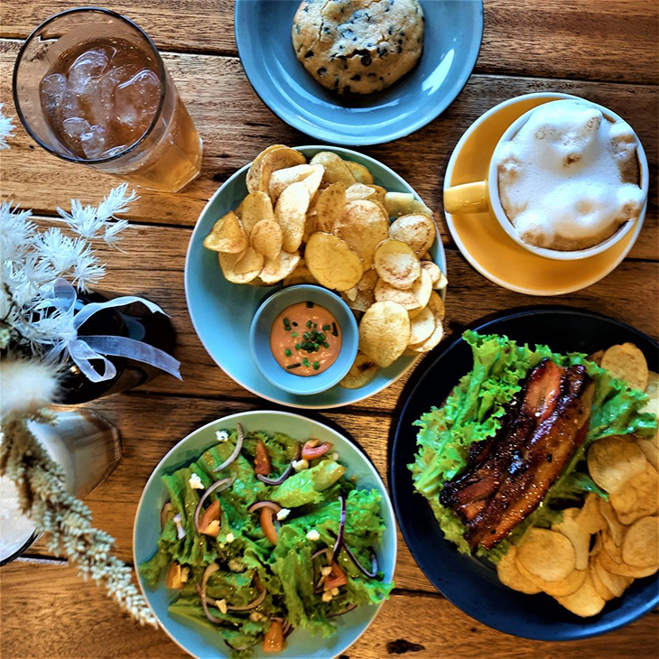 Flat lay of potato chips, salad, bacon, chocolate chip cookie, cappuccino with bear 3D art, and iced tea